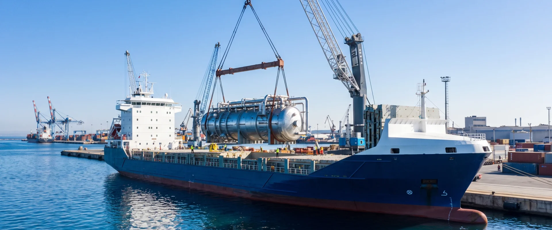 Modern breakbulk cargo vessel with navy blue hull docked at port terminal with heavy-duty crane hoisting oversized industrial module
